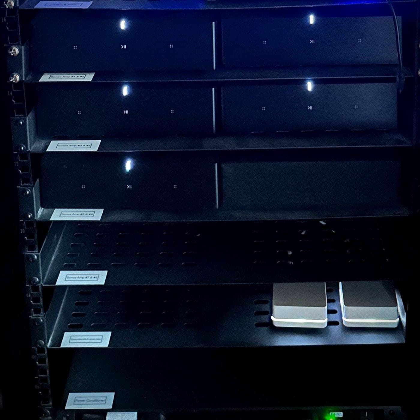 Labelled rack shelves holding pairs of audio amplifiers, each shelf tagged with what it powers