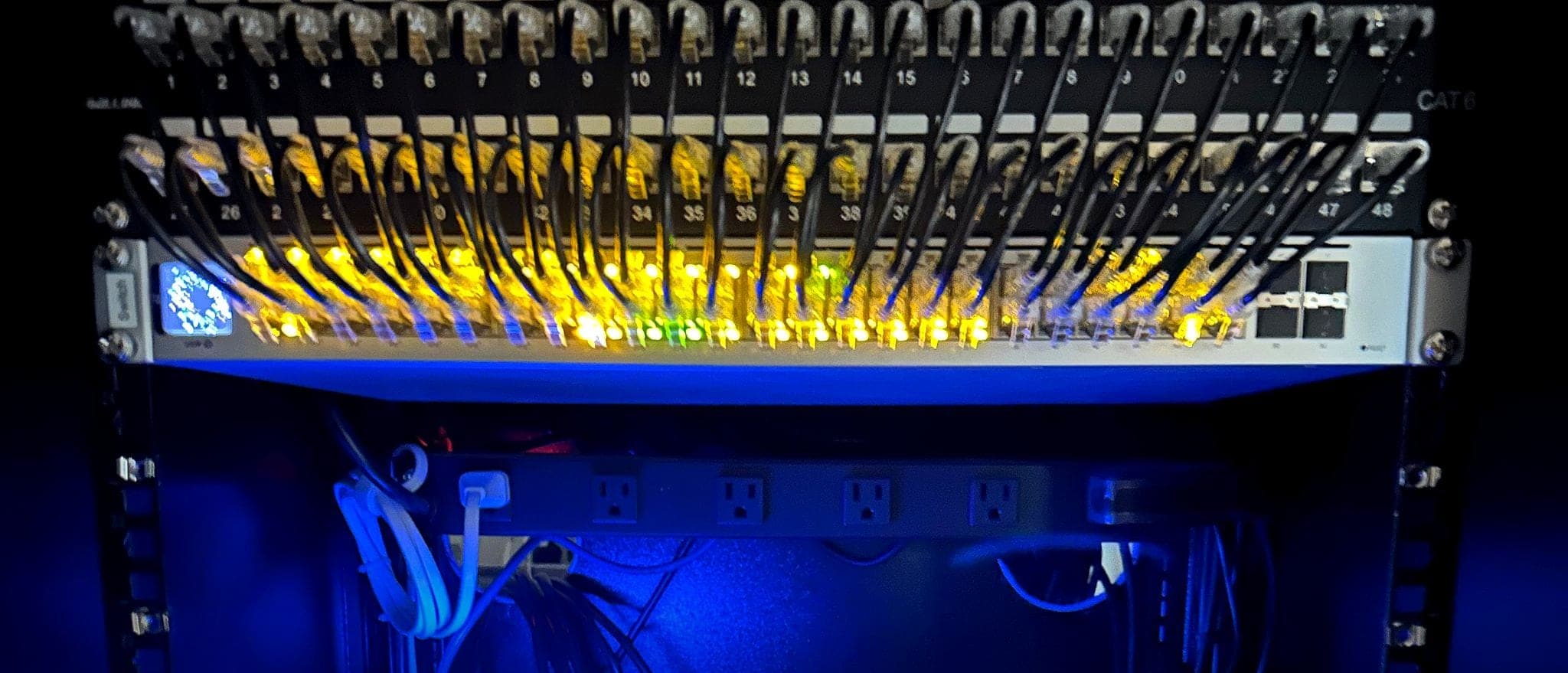 Network rack with the room lights off: two patch panels patched into a 48-port switch with its link lights on, above the gateway and network storage