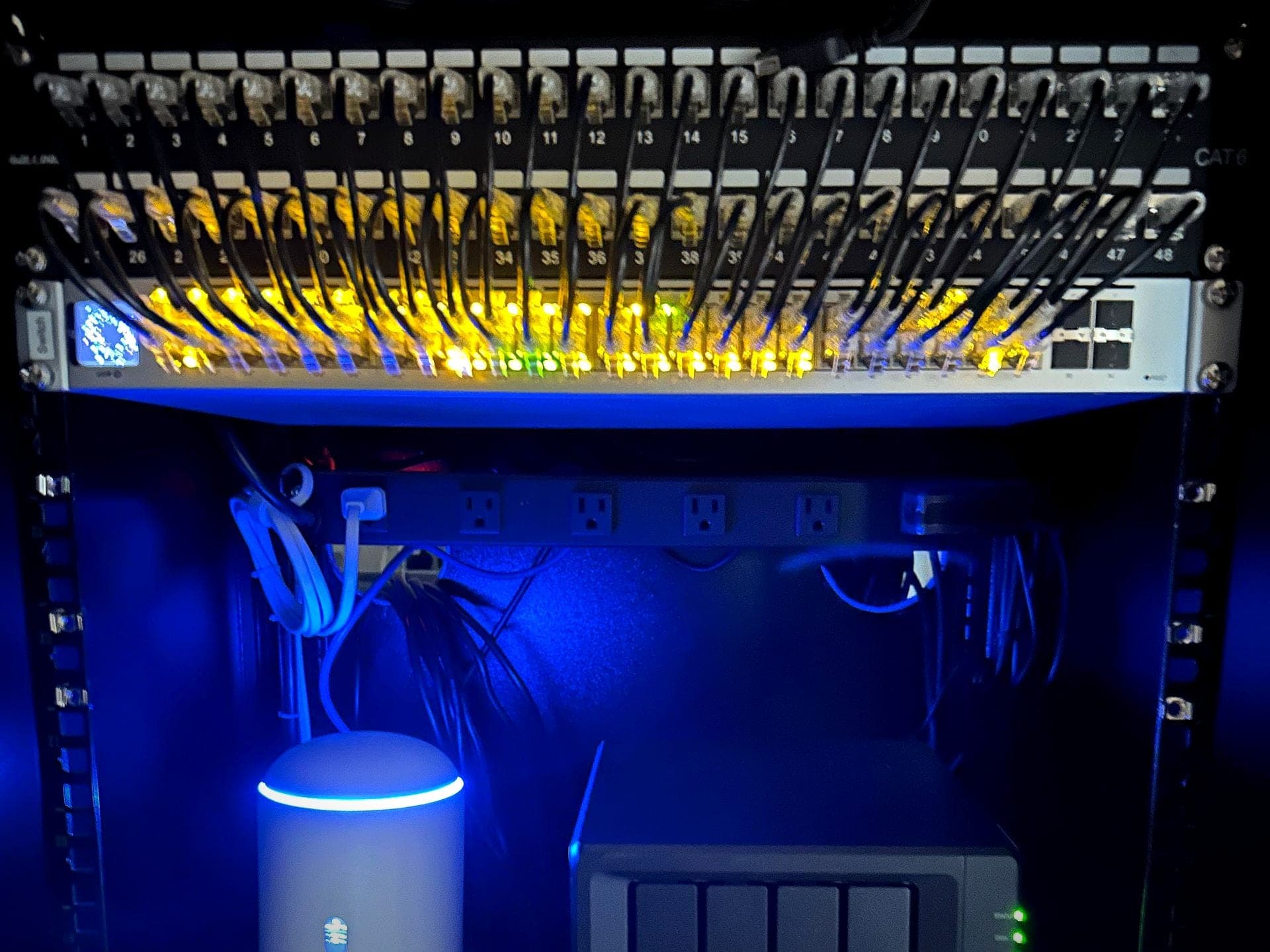Network rack with the room lights off: two patch panels patched into a 48-port switch with its link lights on, above the gateway and network storage